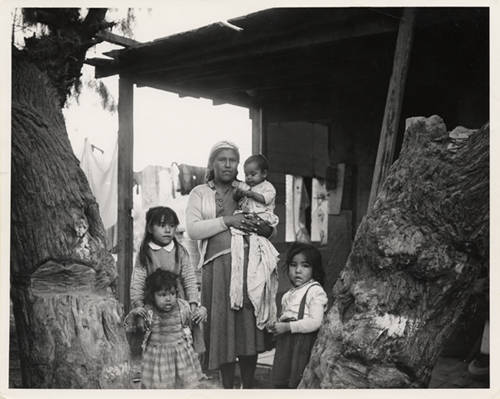 Woman holding a baby surrounded by 3 small children outside a dilapidated building
