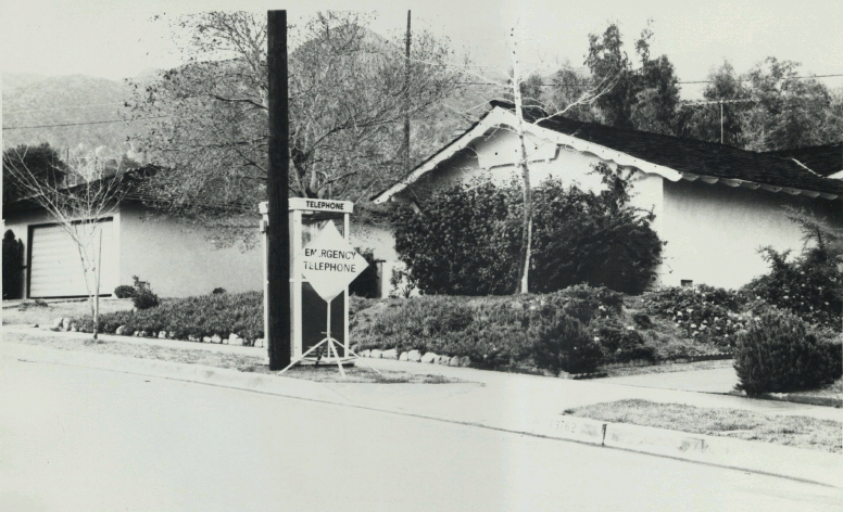 View from the street of a house with a phone booth at the curb. Sign says Emergency Telephone.