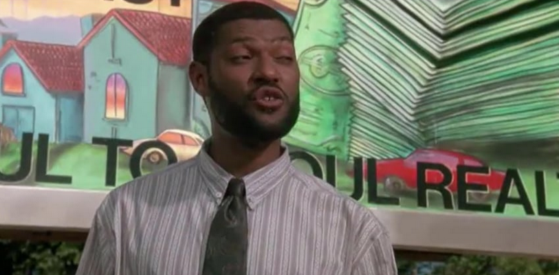 Man speaking to someone not in picture with a billboard in the background; billboard contains drawings of houses and money.