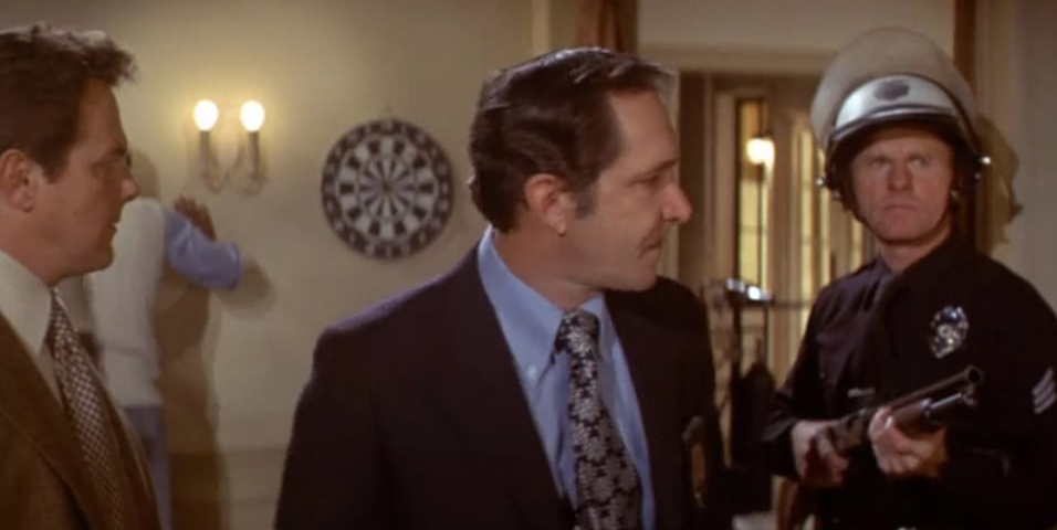 Three men, one dressed in police uniform, outside of a room. In the background one man is facing the wall with hands on the wall while being frisked.